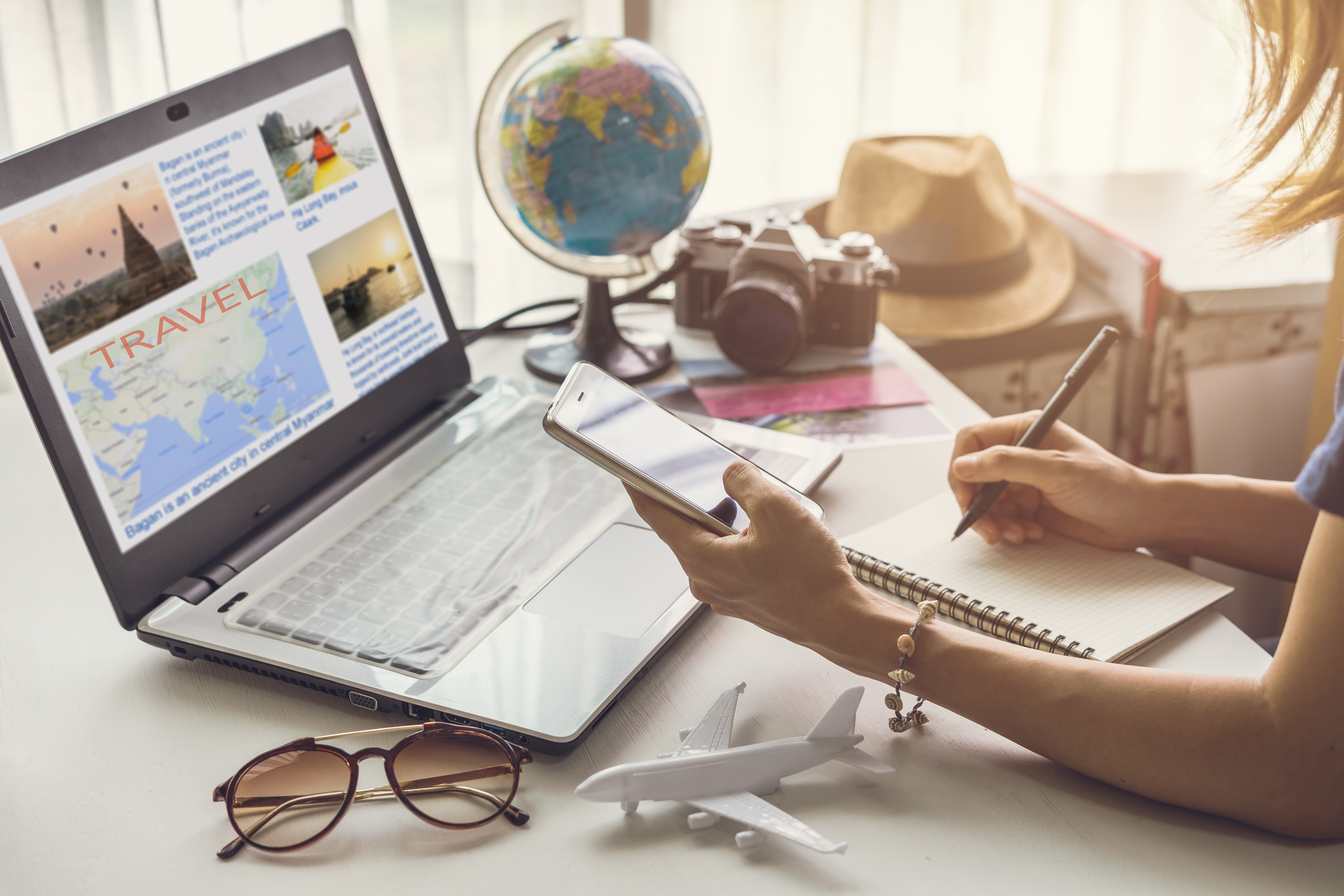 image of someone planning on how to save for vacation; laptop in view with camera, hat, phone, and hand with pen